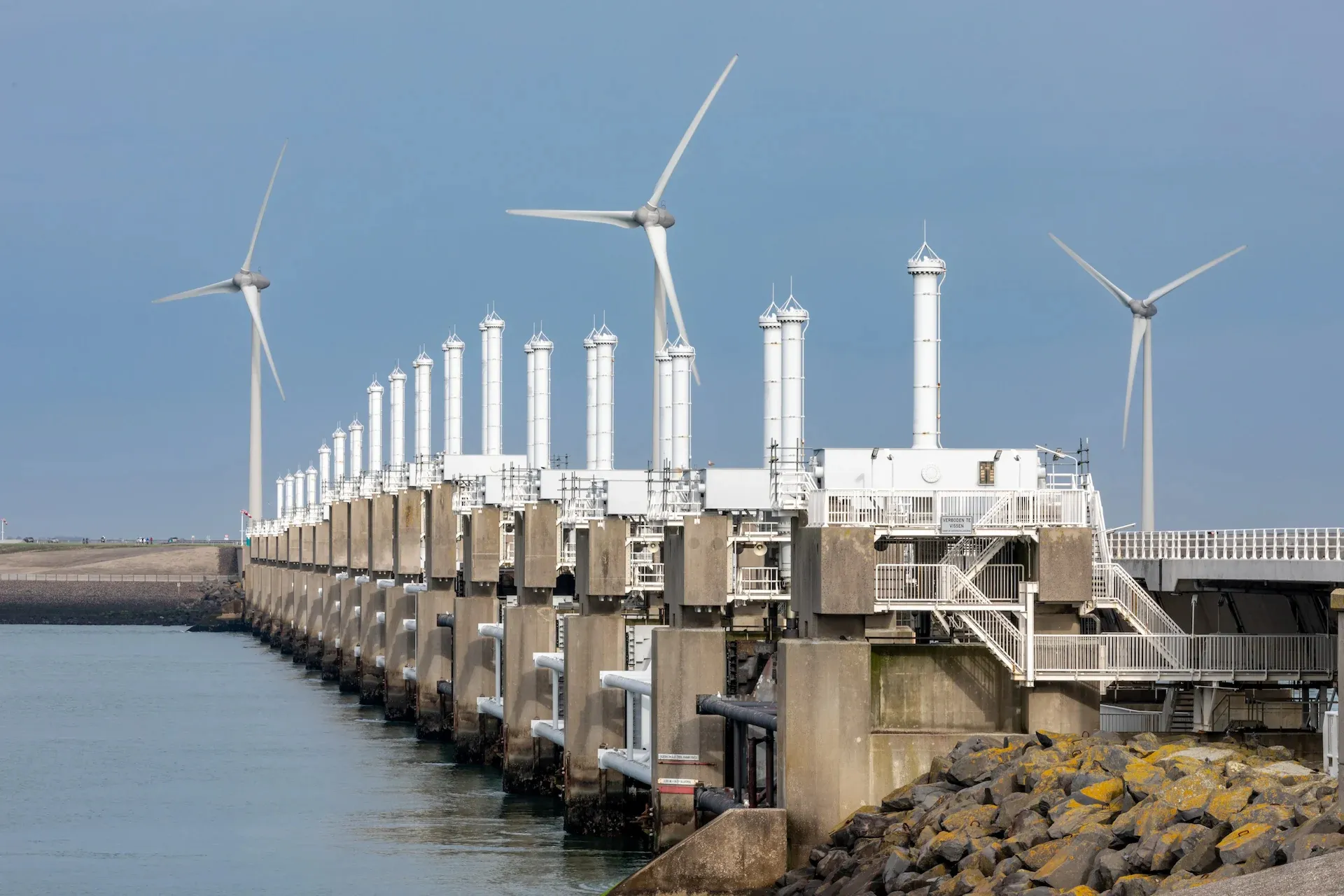 Aerial view of the Oosterscheldekering storm surge barrier, a long line of concrete piers and sluice gates spanning the Eastern Scheldt estuary, with wind turbines standing along its length.