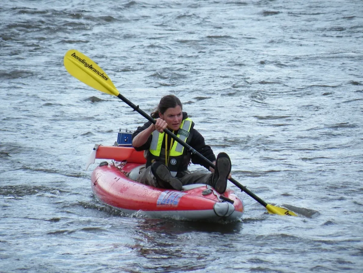 USGS field staff carrying flow measuring equipment across Ikalukrok Creek, a shallow river in a remote Alaskan landscape.