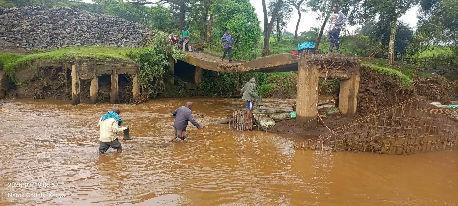 A river in spate carrying debris where a road bridge has been washed away, its broken deck left hanging over the water.